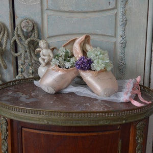 May include: A pair of pink ballet pointe shoes filled with white and purple flowers, resting on a marble-topped table. A small cherub statue and a pink and white ribbon are also on the table. The background features antique doors.