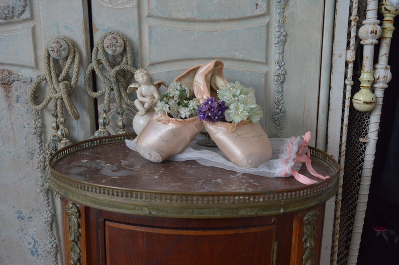 May include: A pair of pink ballet pointe shoes filled with white and purple flowers, resting on a marble-topped table. A small cherub statue and a pink and white ribbon are also on the table. The background features antique doors.