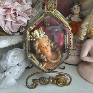 May include: A vintage, hexagonal glass and brass display case with a doll head wearing a crown. The case is on a decorative brass stand. Other dolls and pink and white floral accents are in the background.