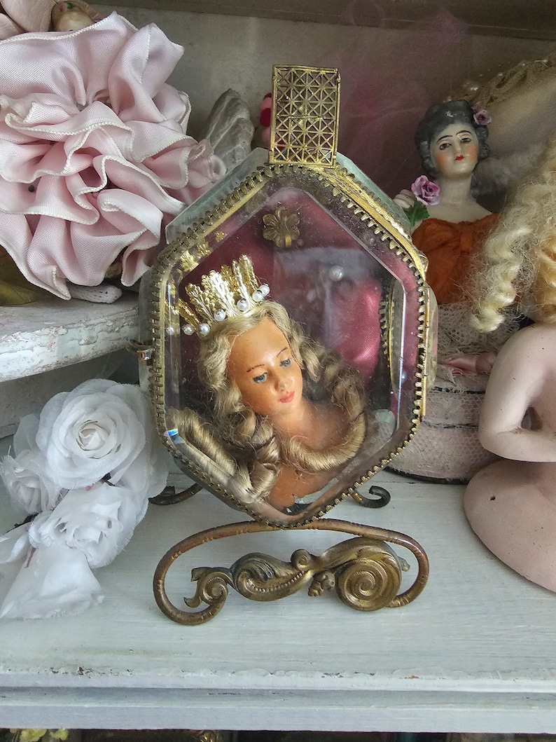 May include: A vintage, hexagonal glass and brass display case with a doll head wearing a crown. The case is on a decorative brass stand. Other dolls and pink and white floral accents are in the background.