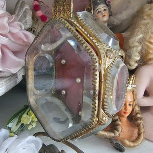 May include: A vintage, clear glass and gold-toned metal jewelry box with a hinged lid. The box has a burgundy interior with small pearls. The box is supported by an ornate, curved metal stand. Doll heads are in the background.