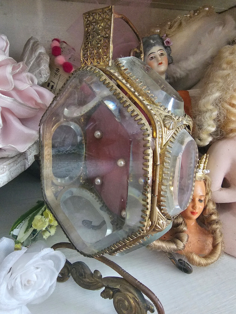 May include: A vintage, clear glass and gold-toned metal jewelry box with a hinged lid. The box has a burgundy interior with small pearls. The box is supported by an ornate, curved metal stand. Doll heads are in the background.