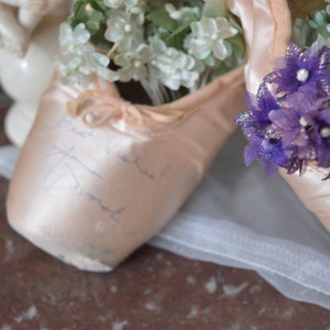 May include: A pair of pink satin ballet pointe shoes, filled with white and purple floral arrangements. The shoes have ribbons and handwritten script. A white tulle fabric is draped beneath the shoes.