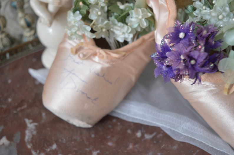 May include: A pair of pink satin ballet pointe shoes, filled with white and purple floral arrangements. The shoes have ribbons and handwritten script. A white tulle fabric is draped beneath the shoes.