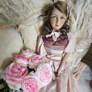May include: A vintage doll with a painted face, wearing a pink dress and a burgundy bodice. The doll is surrounded by cream-colored pillows and a bouquet of pink roses. The background includes lace and fabric.