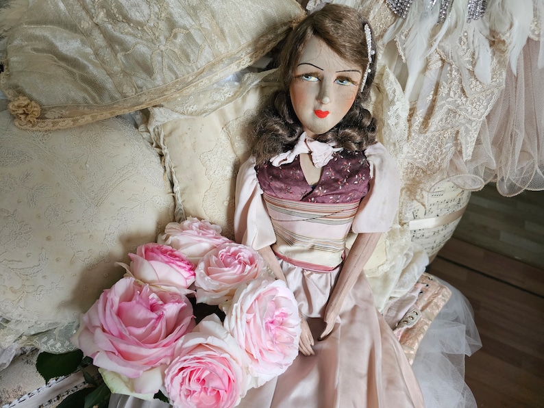 May include: A vintage doll with a painted face, wearing a pink dress and a burgundy bodice. The doll is surrounded by cream-colored pillows and a bouquet of pink roses. The background includes lace and fabric.