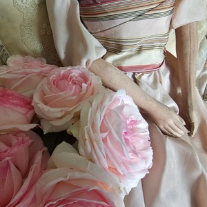 May include: A collection of pale pink roses with darker pink centers, arranged near a doll dressed in a light pink gown with a striped bodice. The doll's arms and hands are visible.