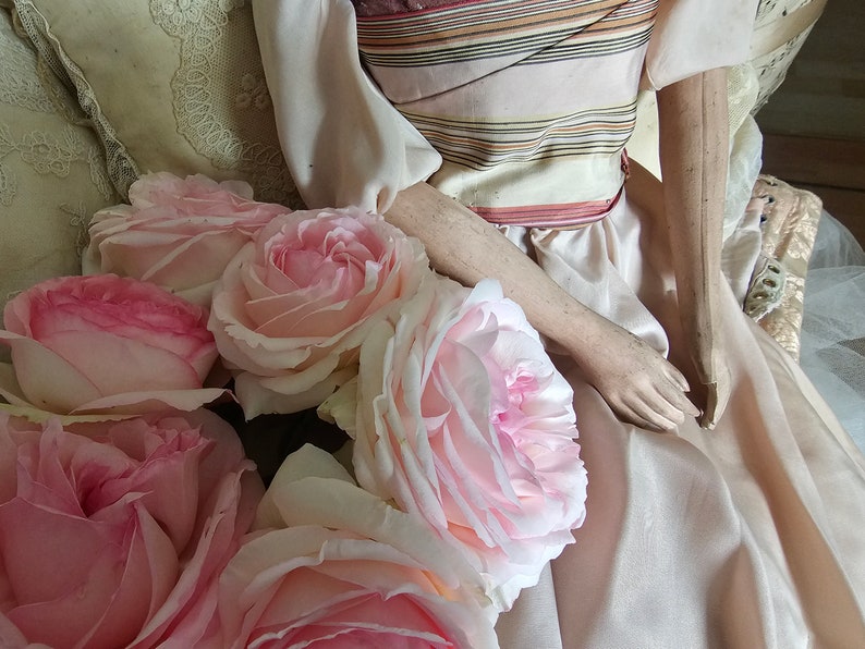 May include: A collection of pale pink roses with darker pink centers, arranged near a doll dressed in a light pink gown with a striped bodice. The doll's arms and hands are visible.
