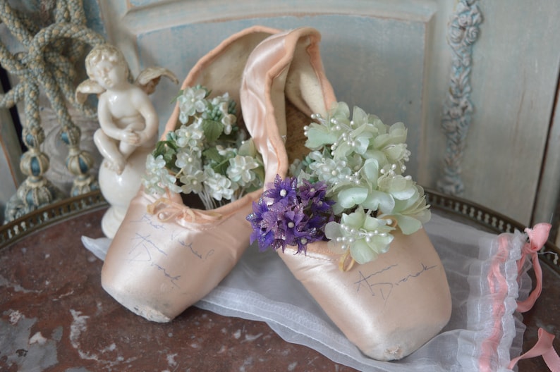 May include: A pair of light pink satin ballet pointe shoes, filled with white and light green flowers, and a small cluster of purple flowers. The shoes have handwritten signatures on them. A decorative cherub statue is in the background.