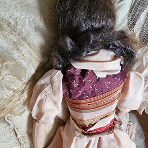 May include: A vintage doll with dark brown hair styled in a low bun. The doll wears a light pink dress with puffed sleeves and a patterned bodice in burgundy and floral print. A striped sash accents the waist.