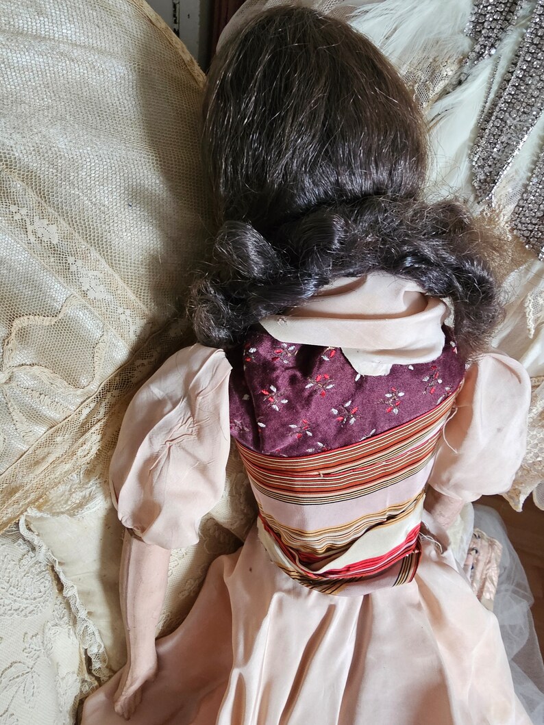 May include: A vintage doll with dark brown hair styled in a low bun. The doll wears a light pink dress with puffed sleeves and a patterned bodice in burgundy and floral print. A striped sash accents the waist.