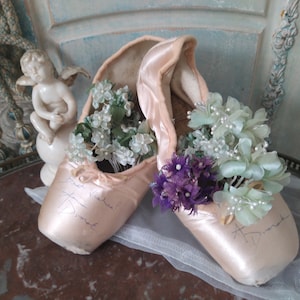 May include: A pair of vintage ballet pointe shoes in a soft pink hue, filled with delicate floral arrangements in shades of white, green, and purple. The shoes have handwritten signatures and are displayed on a decorative surface.