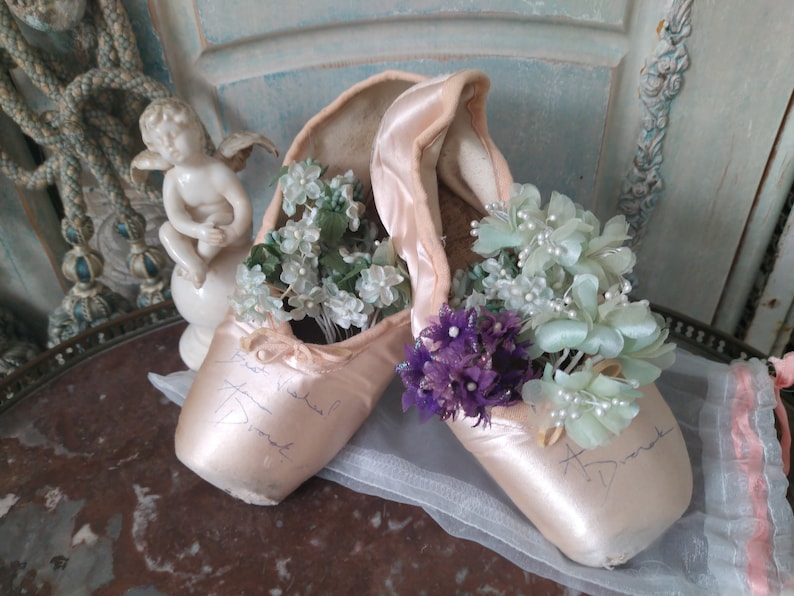 May include: A pair of vintage ballet pointe shoes in a soft pink hue, filled with delicate floral arrangements in shades of white, green, and purple. The shoes have handwritten signatures and are displayed on a decorative surface.