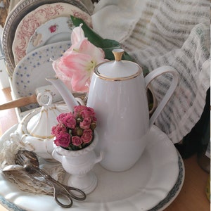 Könnte beinhalten: Eine weiße Porzellan-Kaffeekanne mit Goldrand steht auf einem großen weißen Teller mit gewelltem Rand. Eine kleine weiße Vase enthält einen Strauß rosa Rosen. Vintage-Teller und Besteck sind ebenfalls zu sehen.