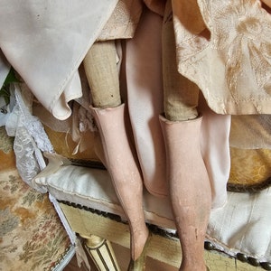 May include: Close-up of an antique doll's legs, showcasing painted pink stockings and olive green heeled shoes. The doll's dress is visible, with layers of cream and peach-colored fabric. The doll is seated on a decorative, antique bench.