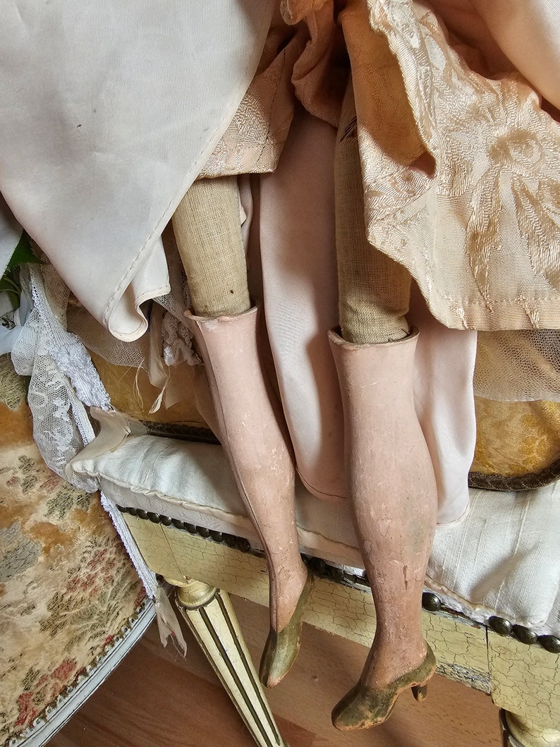 May include: Close-up of an antique doll's legs, showcasing painted pink stockings and olive green heeled shoes. The doll's dress is visible, with layers of cream and peach-colored fabric. The doll is seated on a decorative, antique bench.