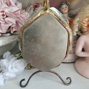 May include: A vintage hand mirror with a hexagonal mirror and a decorative gold-colored frame. The mirror is supported by a curved, ornate stand. The background includes antique dolls and floral decorations.