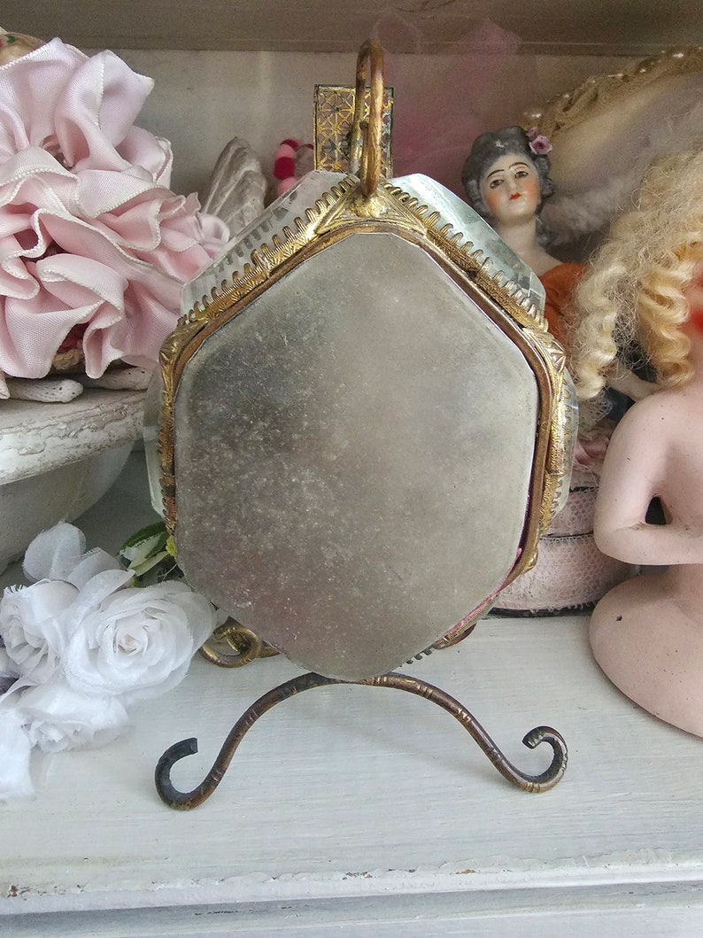 May include: A vintage hand mirror with a hexagonal mirror and a decorative gold-colored frame. The mirror is supported by a curved, ornate stand. The background includes antique dolls and floral decorations.