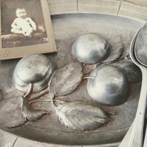 May include: A silver serving dish with a raised design of three plums and leaves. The dish is on a table with a vintage photo of a child in the background.