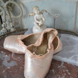May include: A pair of well-worn, light pink satin ballet pointe shoes with visible wear and tear. The shoes are resting on a marble-topped table, alongside a small cherub statue and a decorative metal piece. The background features a light blue wall.