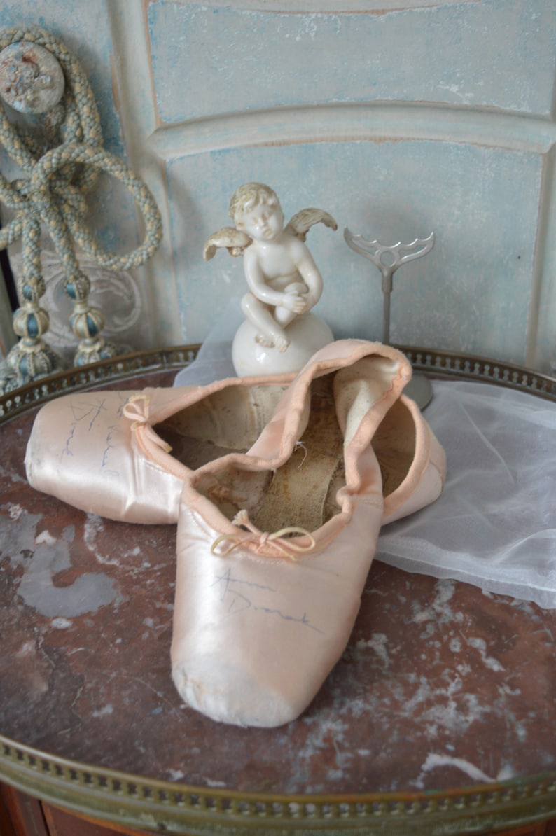 May include: A pair of well-worn, light pink satin ballet pointe shoes with visible wear and tear. The shoes are resting on a marble-topped table, alongside a small cherub statue and a decorative metal piece. The background features a light blue wall.