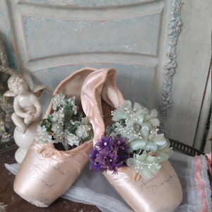 May include: A pair of satin ballet pointe shoes in a soft pink hue, filled with delicate white and purple flowers. The shoes have handwritten signatures and are displayed on a reflective surface.