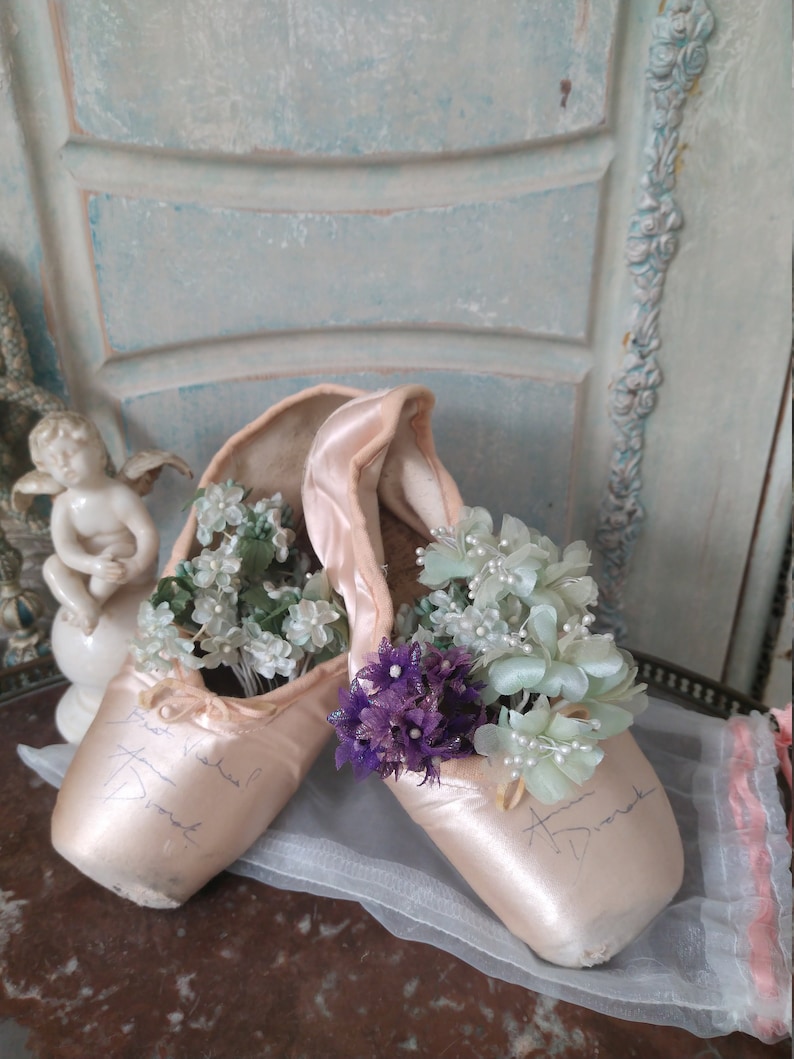 May include: A pair of satin ballet pointe shoes in a soft pink hue, filled with delicate white and purple flowers. The shoes have handwritten signatures and are displayed on a reflective surface.