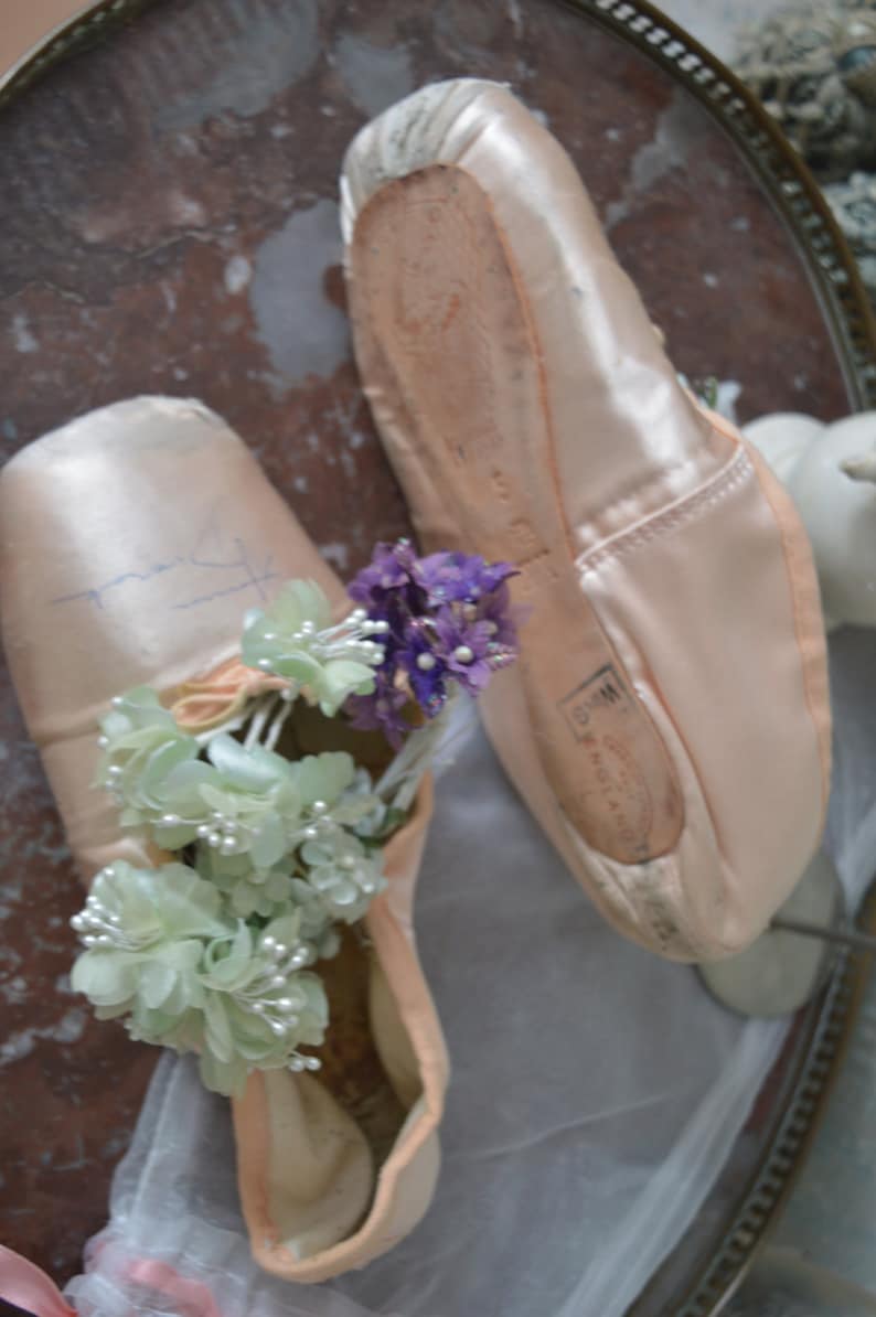 May include: A pair of well-worn, light pink satin ballet pointe shoes. One shoe is decorated with a cluster of light green and white flowers, and the other with purple flowers. The shoes are resting on a decorative surface.