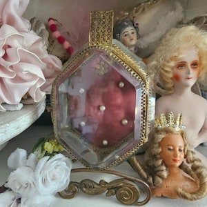 May include: A vintage perfume bottle with a gold filigree top and a burgundy interior, adorned with small pearls. The bottle is set on a decorative stand, surrounded by antique dolls and floral accents, creating a romantic, nostalgic aesthetic.