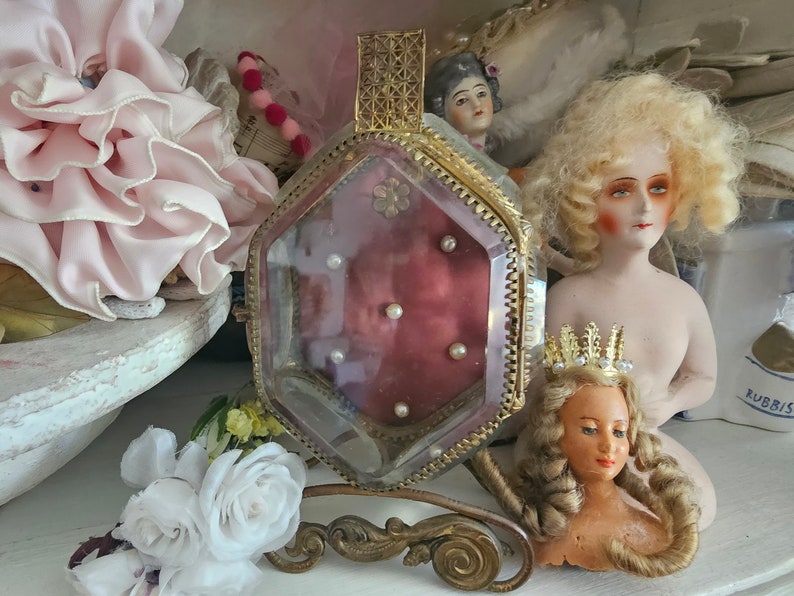 May include: A vintage perfume bottle with a gold filigree top and a burgundy interior, adorned with small pearls. The bottle is set on a decorative stand, surrounded by antique dolls and floral accents, creating a romantic, nostalgic aesthetic.