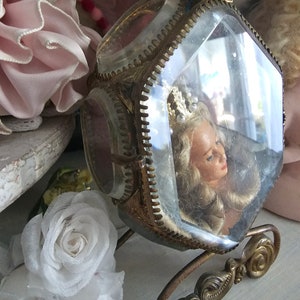 May include: A vintage, ornate, glass and brass carriage-shaped display case. Inside, a doll's head with blonde hair and a crown is visible. The carriage sits on a decorative brass base, with floral accents and a geometric top.