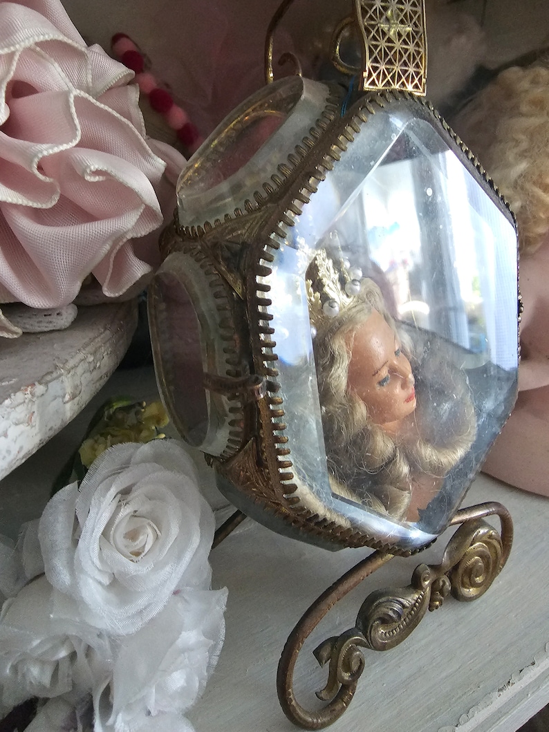 May include: A vintage, ornate, glass and brass carriage-shaped display case. Inside, a doll's head with blonde hair and a crown is visible. The carriage sits on a decorative brass base, with floral accents and a geometric top.