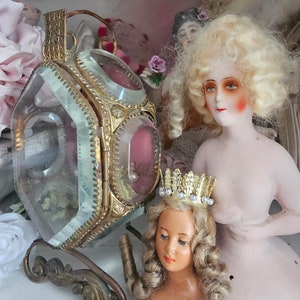 May include: An antique display featuring a gold-toned, glass-paneled lantern with a decorative base, and two vintage porcelain dolls. One doll has blonde curly hair and rosy cheeks, while the other wears a gold crown.