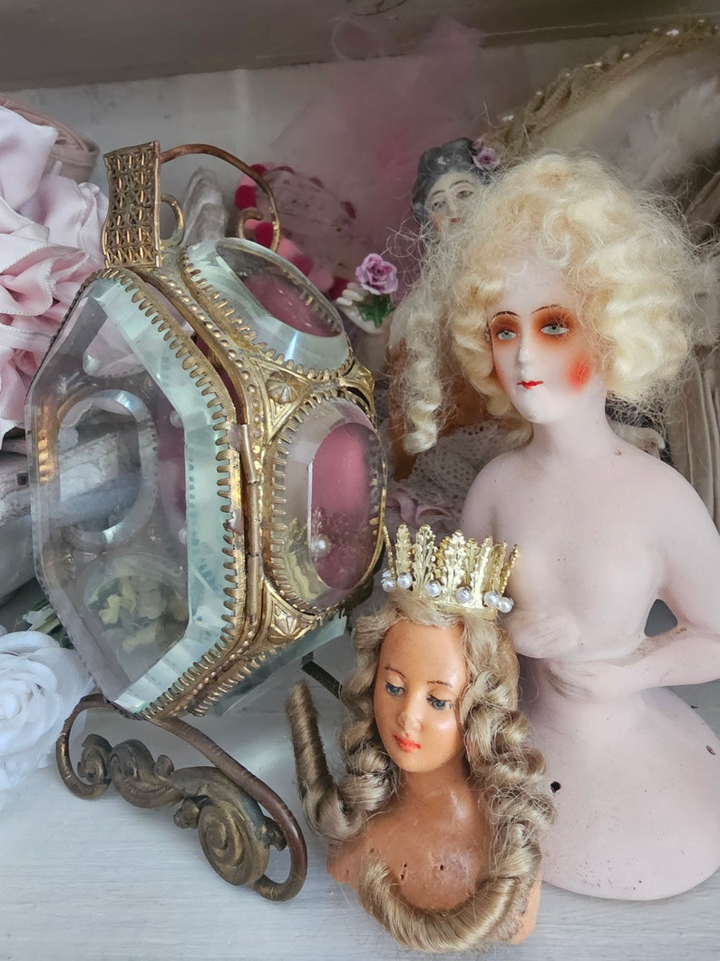 May include: An antique display featuring a gold-toned, glass-paneled lantern with a decorative base, and two vintage porcelain dolls. One doll has blonde curly hair and rosy cheeks, while the other wears a gold crown.