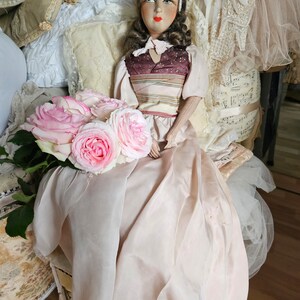 May include: A vintage doll with a painted face, wearing a pink dress and holding a bouquet of pink roses. The doll is seated on a small chair with a cream-colored cushion. The background includes pillows and vintage clothing.