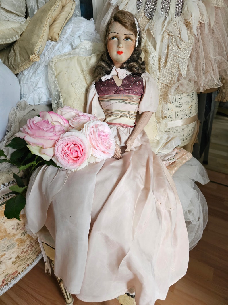May include: A vintage doll with a painted face, wearing a pink dress and holding a bouquet of pink roses. The doll is seated on a small chair with a cream-colored cushion. The background includes pillows and vintage clothing.
