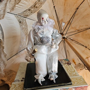 May include: A porcelain clown doll wearing a white and silver costume, sitting on a black base with a gold trim. The doll is holding a pair of drumsticks and has a painted face with red cheeks and a single red tear.