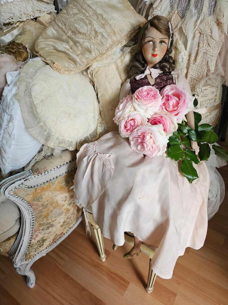 May include: A vintage doll with painted features, wearing a pink dress and holding a bouquet of pink roses. The doll is seated on a small chair with cream-colored legs. Pillows and other vintage items surround the doll.