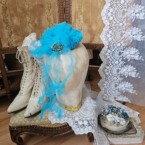 May include: A vintage display featuring a turquoise fascinator with a jeweled brooch, a pair of cream-colored lace-up boots, and a lace trim. The items are arranged on a small upholstered stool.
