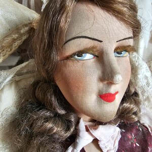 May include: Close-up of a vintage doll's face. The doll has light brown hair, blue eyes, and red lips. The doll is wearing a burgundy vest with a floral pattern and a white ruffled collar. The doll's face is made of fabric.