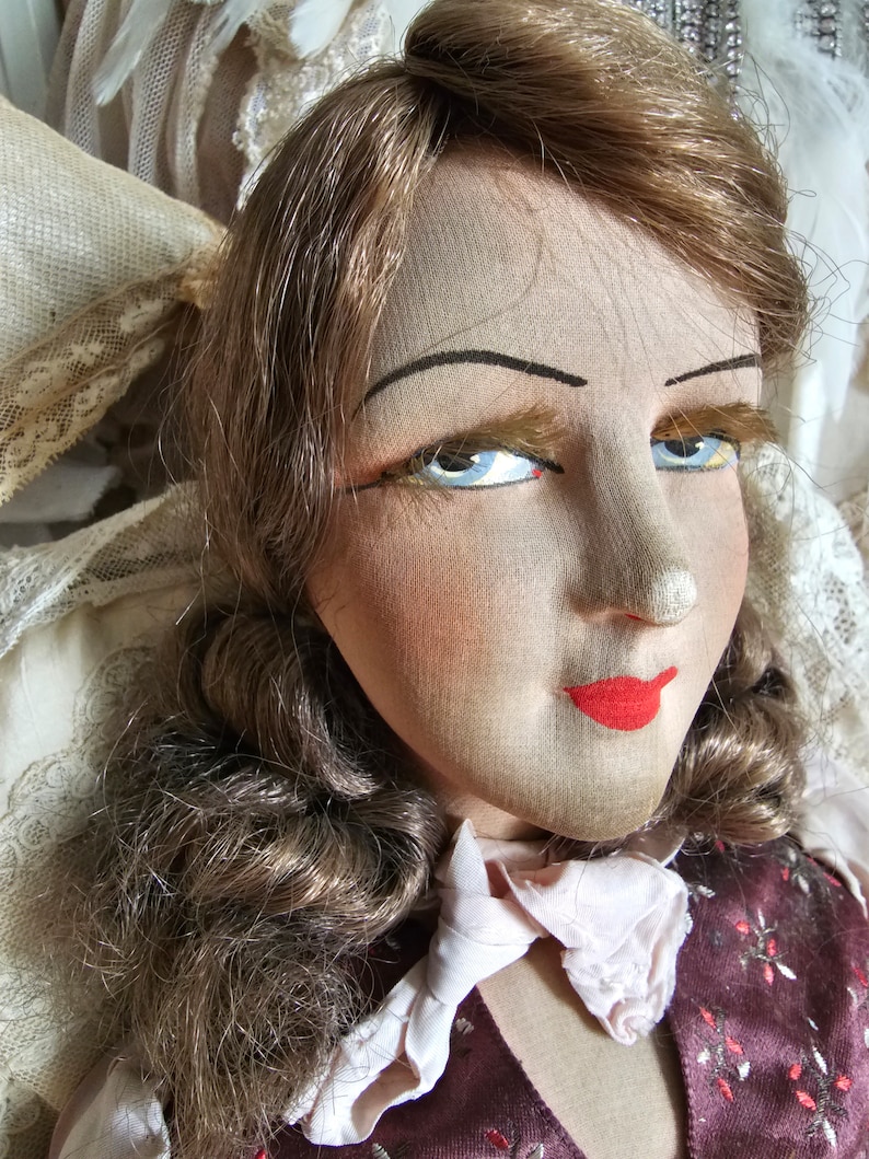May include: Close-up of a vintage doll's face. The doll has light brown hair, blue eyes, and red lips. The doll is wearing a burgundy vest with a floral pattern and a white ruffled collar. The doll's face is made of fabric.