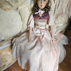 May include: A vintage doll with a painted face, wearing a light pink dress and a burgundy bodice with a striped design. The doll has brown hair and is seated on a decorative chair with pillows in the background.