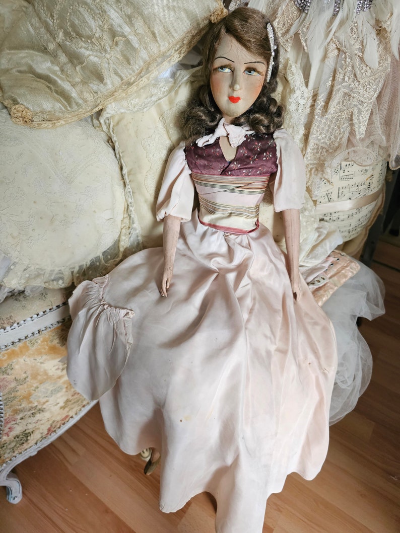 May include: A vintage doll with a painted face, wearing a light pink dress and a burgundy bodice with a striped design. The doll has brown hair and is seated on a decorative chair with pillows in the background.