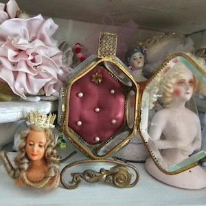 May include: An antique display featuring a small doll with a gold crown, a decorative hinged box with a red interior and pearls, and a doll inside a glass case. The items are arranged on a white surface.