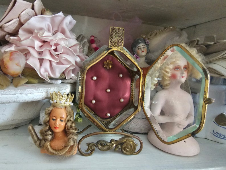 May include: An antique display featuring a small doll with a gold crown, a decorative hinged box with a red interior and pearls, and a doll inside a glass case. The items are arranged on a white surface.