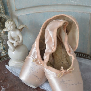 May include: A pair of well-worn, light pink satin ballet pointe shoes with visible wear and tear. Each shoe has a small bow and handwritten text. A small white cherub statue sits nearby, suggesting a vintage or antique theme.