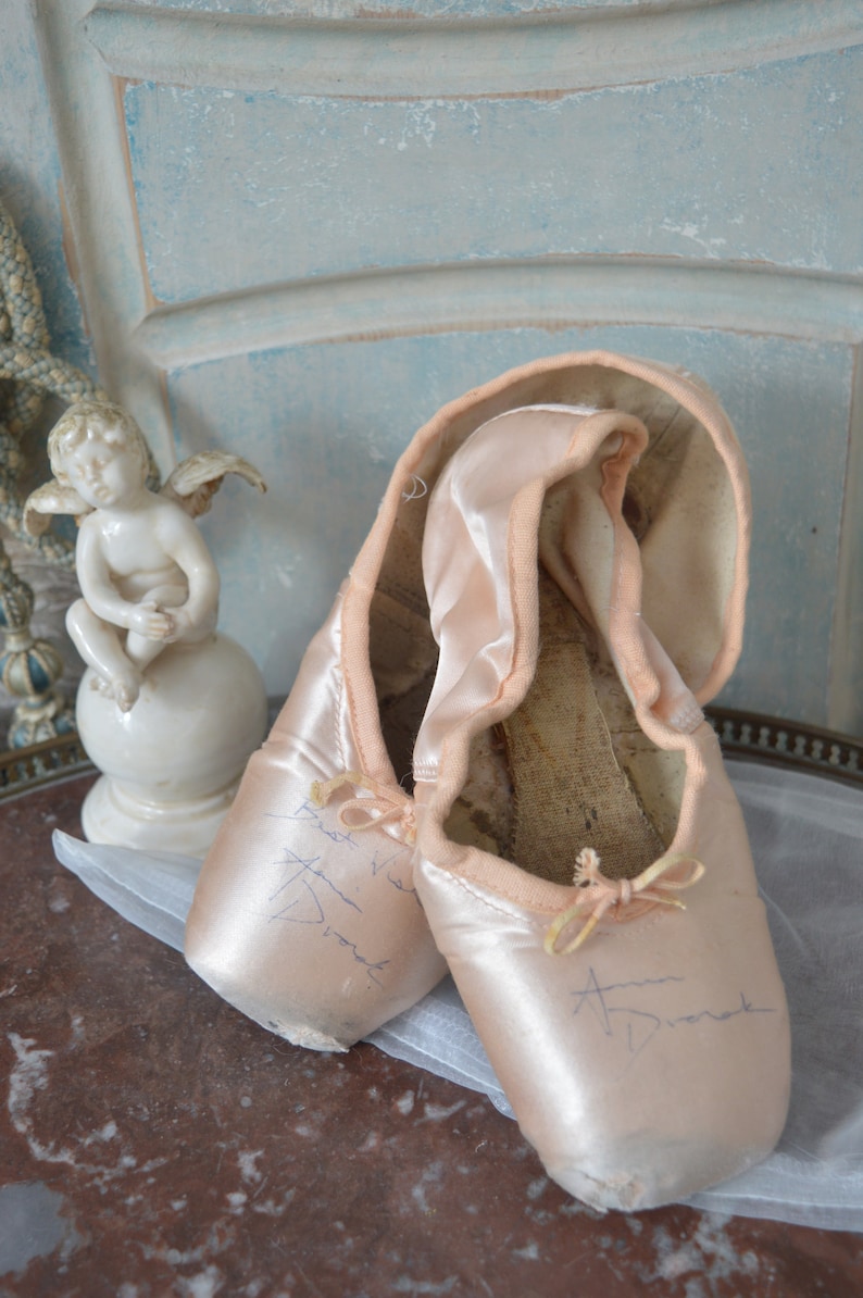 May include: A pair of well-worn, light pink satin ballet pointe shoes with visible wear and tear. Each shoe has a small bow and handwritten text. A small white cherub statue sits nearby, suggesting a vintage or antique theme.