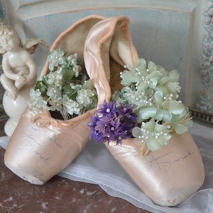 May include: A pair of light pink satin ballet pointe shoes, filled with white and light green flowers, and a small cluster of purple flowers. The shoes have handwritten signatures on them. A decorative cherub statue is in the background.
