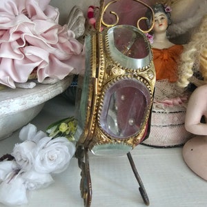 May include: An antique, ornate, gold-toned metal and glass vanity mirror with a stand. The mirror has a rectangular shape with rounded corners and is decorated with intricate detailing. The glass is clear with a pinkish hue. The mirror is displayed on a white shelf.