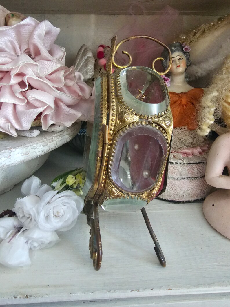 May include: An antique, ornate, gold-toned metal and glass vanity mirror with a stand. The mirror has a rectangular shape with rounded corners and is decorated with intricate detailing. The glass is clear with a pinkish hue. The mirror is displayed on a white shelf.